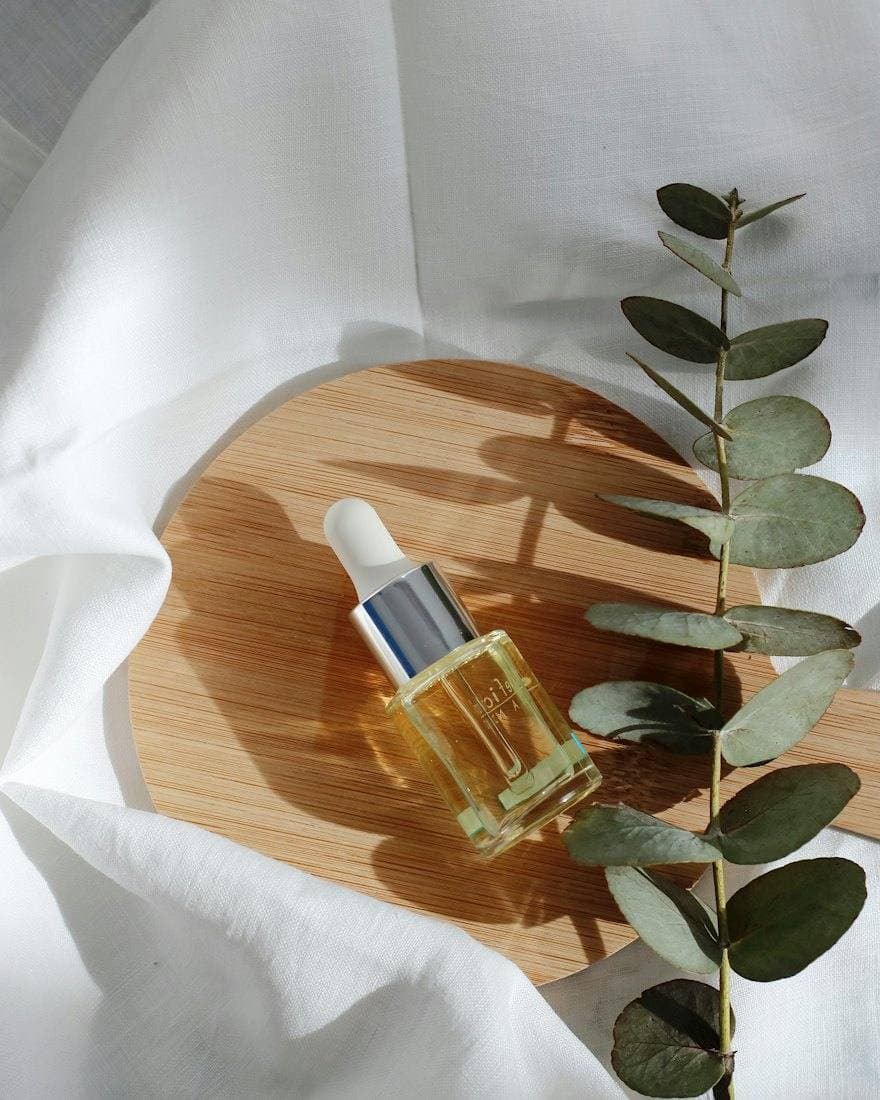 A facial oil with a dropper resting on a wooden board beside eucalyptus
